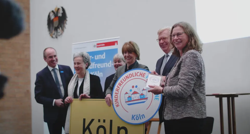 Menschen bei einer Veranstaltung in Köln für eine kinderfreundliche Kommune.  Ein Schild zeigt das Logo 'Kinderfreundliche Kommune' mit Häusern und Kindern.  Ein Kölner Stadtschild ist ebenfalls zu sehen.  Der Hintergrund zeigt das deutsche Bundeswappen und ein Banner der Stadt Köln.