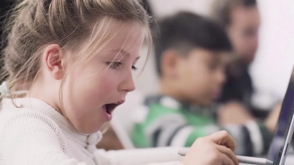 Junges Mädchen, überrascht beim Arbeiten mit einem Tablet im Klassenzimmer.  Andere Schüler im Hintergrund.