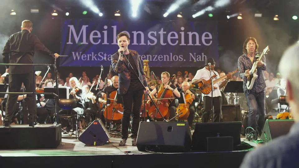 Live-Musikperformance mit Sänger, Gitarrist und Band, begleitet von Orchester und Chor. Beleuchtete Bühne mit Banner: „Meilensteine WerkStatt. Grenzenlos. Nah.“